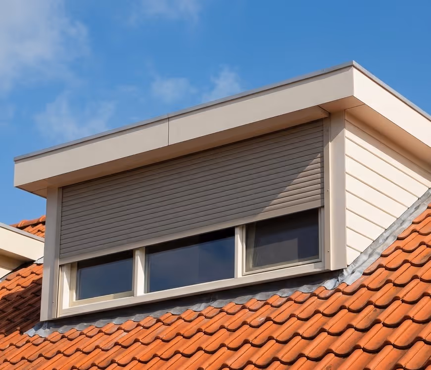 Moderne dakkapel met deels gesloten rolluik op een dak met rode dakpannen onder een blauwe lucht.